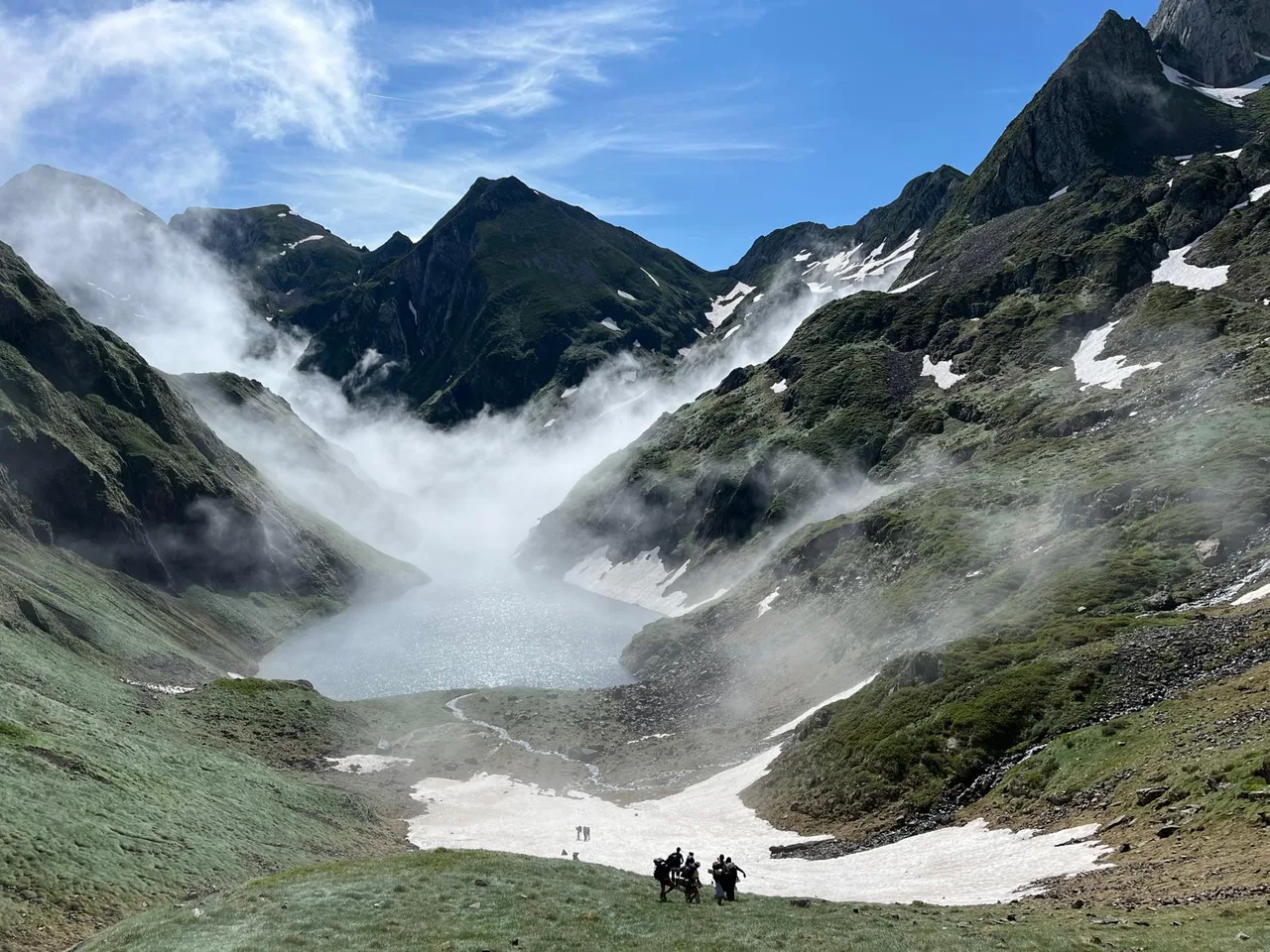 Un estany entre cims, neu i boira amb excursionistes d’esquena al fons